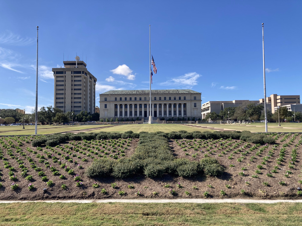 The US and Texan flags, half mast on a flagpole in the middle of a field, in front of a building.