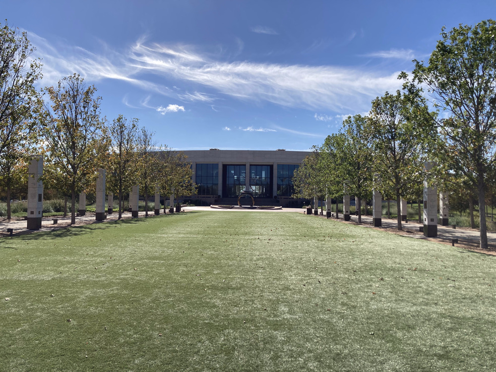 A building with a curved wall and large windows is visible, across a lawn from the viewer. On either sides of the lawn are trees and stone columns.