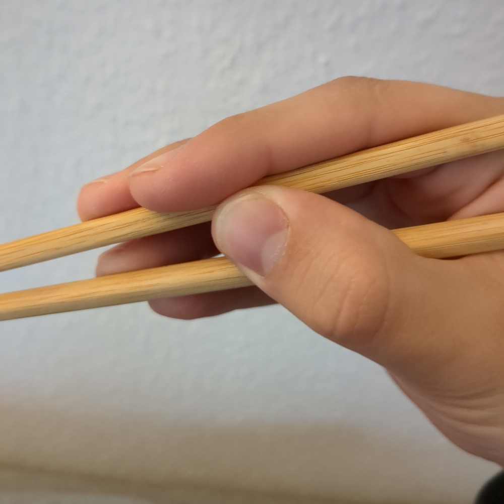 Chopsticks held incorrectly, with the index and middle finger touching the upper stick and the ring and pinky finger holding the lower stick.