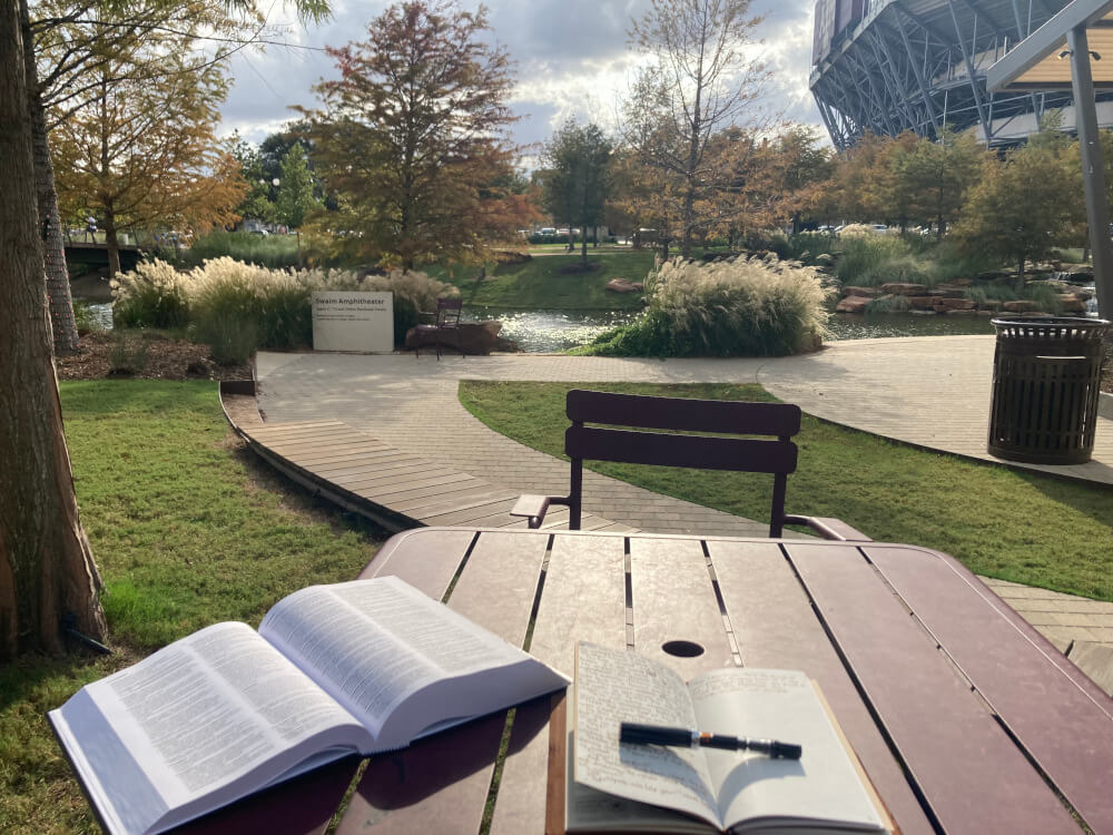A table in the park, overlooking a pond. On the table is a notebook, capped fountain pen, and thesaurus.
