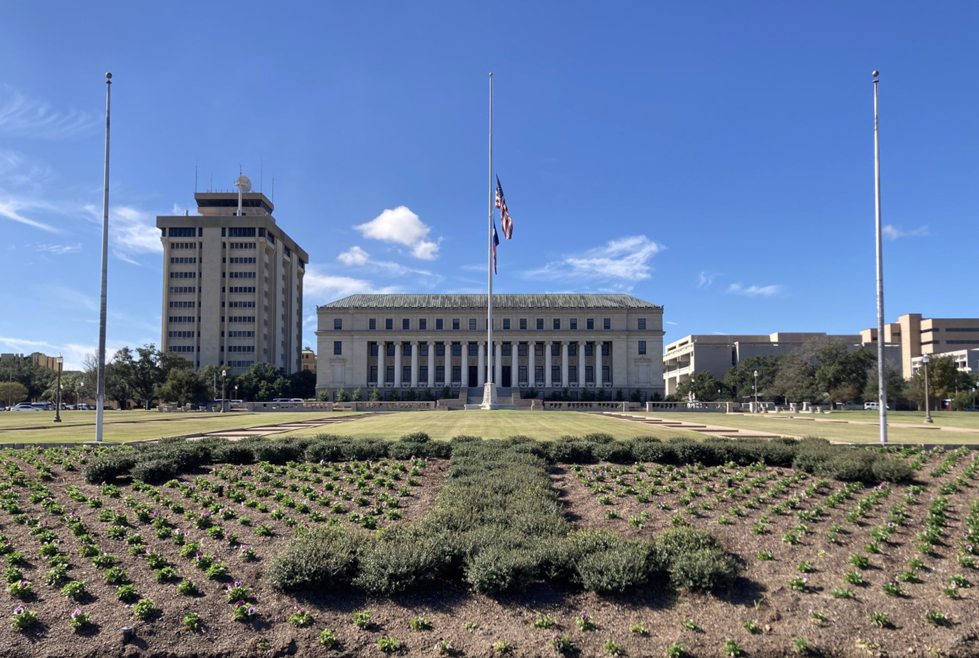 The US and Texan flags, half mast on a flagpole in the middle of a field, in front of a building.